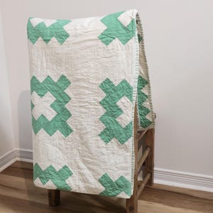 Op de afbeelding: Een handgemaakte quilt met een witte achtergrond en een herhalend patroon van groene kruisen. De quilt is over een houten stoel gedrapeerd, waarbij het ontwerp en de textuur worden getoond. De zichtbare stiksels benadrukken het vakmanschap.