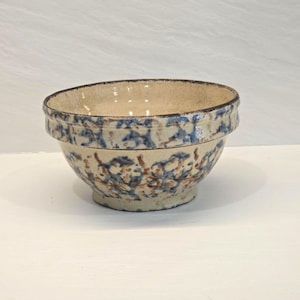 Puede incluir: Un cuenco de cerámica con una base de color crema que presenta un patrón de esponjado en azul y marrón. El cuenco tiene un borde horizontal distintivo cerca de la parte superior y un borde oscuro y texturizado alrededor de la abertura.