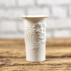 May include: A matte white ceramic vase featuring a flared rim and a relief design of sculpted flowers with long, sweeping petals extending down the side. The surface is smooth and uniform in color, showcasing intricate floral detailing.
