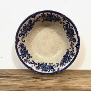 May include: A round ceramic bowl featuring a cream-colored center and a wide, dark blue decorative border. The rim is adorned with intricate floral patterns and scrolling foliage designs that encircle the entire edge of the bowl.