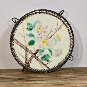 Può includere: Un piatto in ceramica vintage con un motivo floreale di fiori rosa e gialli su sfondo bianco. Il piatto è incorniciato da un supporto in filo metallico nero.