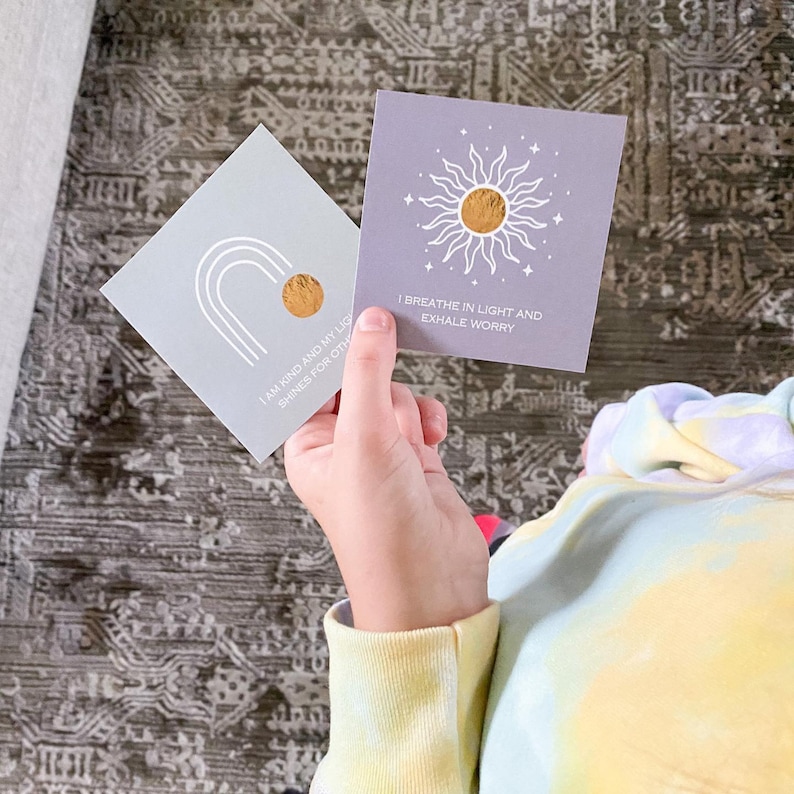 May include: Two grey affirmation cards with gold foil accents. The first card features a rainbow arch and the text "I am kind and my light shines for others". The second card features a sun with the text "I breathe in light and exhale worry".