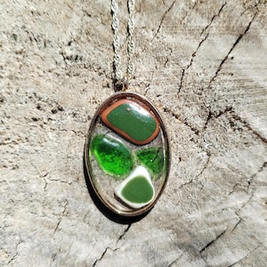 May include: Oval pendant necklace with a silver chain. The pendant features green and white stones set in a clear resin with a gold-coloured border. The background is a weathered wood surface.