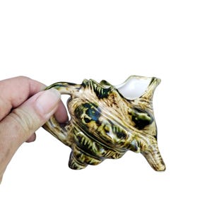 May include: A ceramic seashell creamer with a handle and spout. The creamer is painted in shades of green, brown, and beige, with a glossy finish. The creamer is approximately 10 cm long and 8 cm tall.