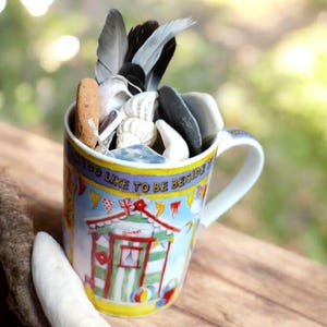 May include: A decorated ceramic mug filled with shells, feathers, and pebbles. The mug has a beach hut design and the words "I DO LIKE TO BE BESIDE" around the rim. The mug is on a wooden surface.