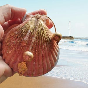 May include: A pink seashell with gold accents and a gold tassel hanging from it. The seashell is being held in a hand with the beach in the background.