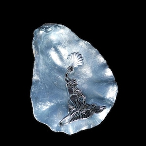 May include: A silver mermaid sculpture holding a fan, set in a light blue, shell-shaped dish. The mermaid has flowing hair and a gown. The shell has a textured surface. The item is set against a black background.