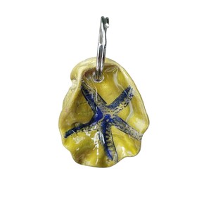 May include: A handmade keychain featuring a yellow seashell with a blue starfish design. The keychain has a silver-coloured metal ring. The seashell is approximately 5 cm in length.
