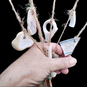 May include: A hand holds a collection of white ceramic charms with various shapes and designs, including a ring, a heart, and a teardrop. The charms are attached to twine and are hanging from the hand.