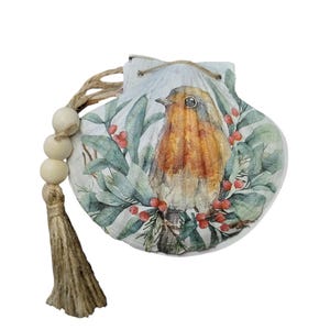 May include: A white seashell with a painted image of a robin perched on a branch with red berries. The seashell has a jute tassel and a string of wooden beads.