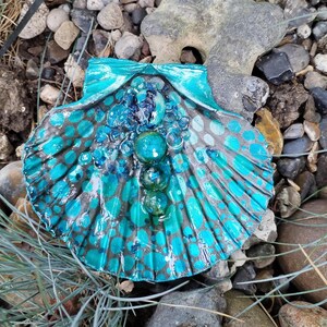 May include: A teal and grey painted decorative seashell, embellished with teal glass beads. The shell has a dotted pattern and a glossy finish, giving it a textured look. This unique seashell is a piece of art.