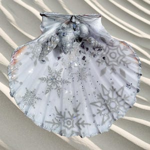 May include: A decorative scallop shell featuring a winter theme. The white shell is adorned with grey snowflake patterns and silver glitter. The shell's edges are outlined in silver. It rests on a sandy surface.
