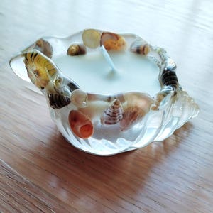 May include: A clear resin candle holder shaped like a seashell, with various seashells embedded within. It holds a white candle with a wick. The candle holder is on a wooden surface.