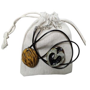 May include: Two necklaces with pendants, one a walnut shell and the other a heart-shaped design, are displayed on a small drawstring bag. The walnut pendant is brown and yellow, while the heart pendant is black and grey. The necklaces have black cords.