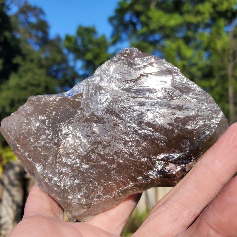 May include: A large, rough-hewn smoky quartz crystal. The crystal is a translucent, brownish-gray color with a reflective, shimmering surface. The natural stone is held in a hand against a backdrop of green trees and a blue sky.