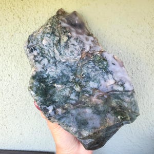 May include: A large, raw moss agate stone with a mix of green, white, and blue hues. The natural stone has a rough, textured surface and is held in a hand.