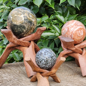 May include: Three decorative stone spheres in shades of green, red, and gray, each displayed on a unique wooden stand. The stands have a natural wood finish and a sculptural design. The spheres vary in size and pattern, creating a natural, earthy aesthetic.