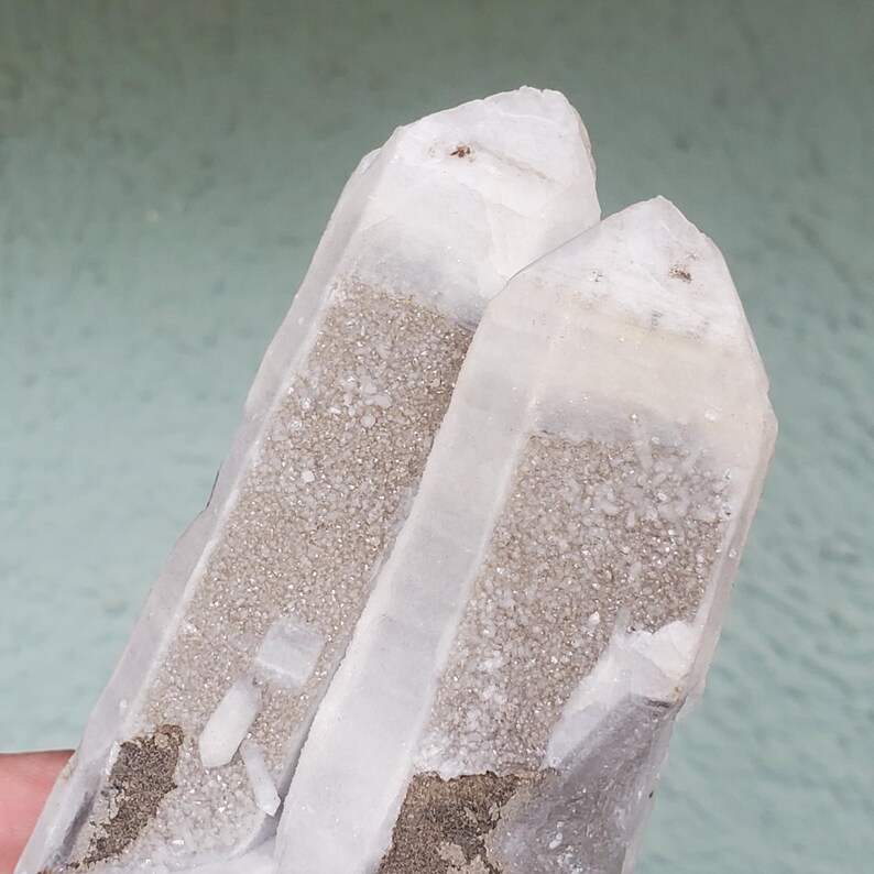 Pu&ograve; includere: Due punte di cristallo di quarzo trasparente con un'inclusione granulare grigio chiaro. I cristalli hanno una consistenza naturale e ruvida e sono parzialmente traslucidi. Il colore generale &egrave; un mix di bianco e grigio, con alcune aree marrone scuro.