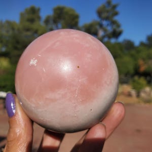 Peut inclure: Une sph&egrave;re en cristal de quartz rose polie, de couleur rose p&acirc;le avec des veines blanches. La boule de cristal est tenue dans une main, avec un arri&egrave;re-plan d'arbres et un ciel bleu. Objet d&eacute;coratif.