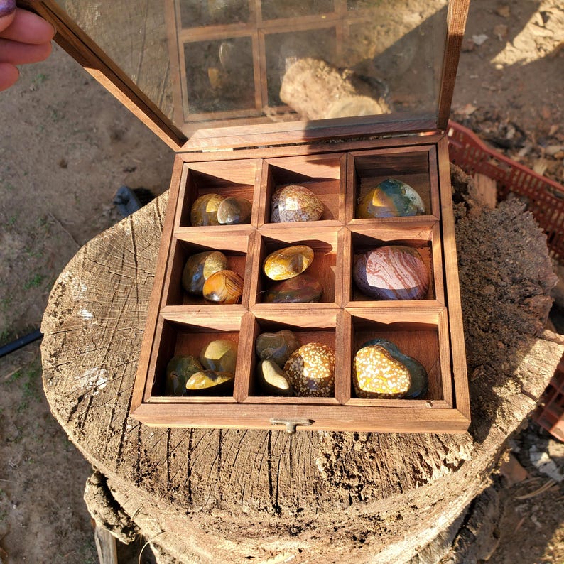 Op de afbeelding: Een houten doos met een glazen deksel, met een verzameling gekleurde stenen. De doos heeft 12 vakken, elk met een andere steen. De stenen zijn in verschillende tinten bruin, rood en wit.