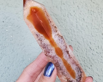 May include: A polished, elongated crystal point featuring a translucent, pale peach outer layer and a deep, vibrant orange core. The center displays a distinct, dark brown, textured mineral inclusion running vertically through the stone.