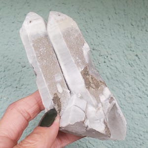 Pu&ograve; includere: Un gruppo di cristalli di quarzo, con una struttura cristallina trasparente e una tonalit&agrave; grigio chiaro e beige. I cristalli sono tenuti in una mano, mostrando la loro consistenza naturale e ruvida e la loro formazione unica. Lo sfondo &egrave; una superficie strutturata verde chiaro.
