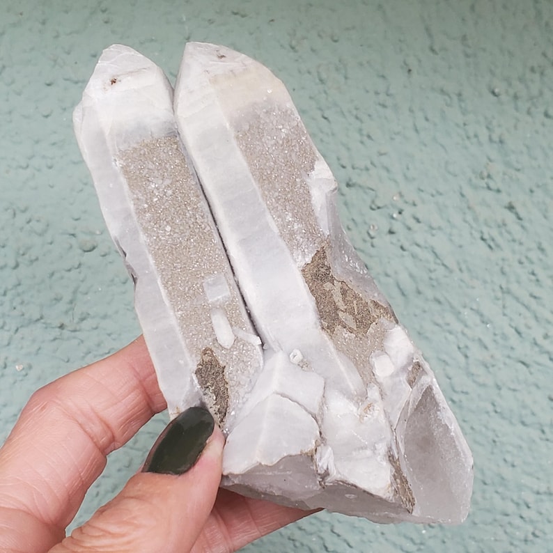 Pu&ograve; includere: Un gruppo di cristalli di quarzo, con una struttura cristallina trasparente e una tonalit&agrave; grigio chiaro e beige. I cristalli sono tenuti in una mano, mostrando la loro consistenza naturale e ruvida e la loro formazione unica. Lo sfondo &egrave; una superficie strutturata verde chiaro.