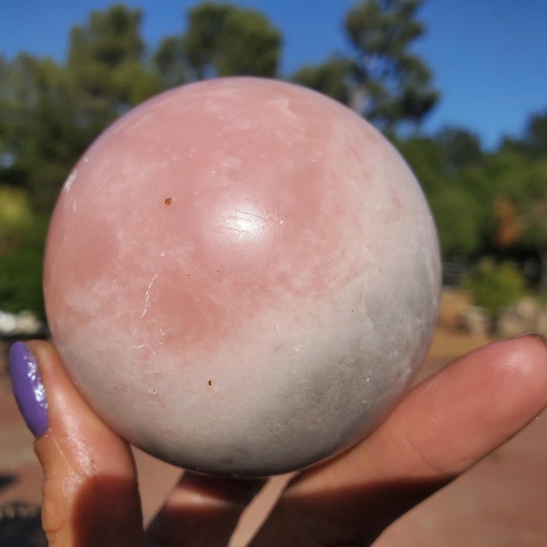Peut inclure: Une sph&egrave;re en quartz rose polie, pr&eacute;sentant un d&eacute;grad&eacute; de rose clair &agrave; blanc et gris. La boule de cristal est tenue dans une main. L'arri&egrave;re-plan montre des arbres et un ciel bleu. Objet d&eacute;coratif.