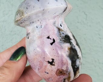 Puede incluir: Una escultura de hongo de piedra pulida en tonos rosa, blanco y negro. El hongo tiene un sombrero redondeado y un tallo liso y curvo. La piedra tiene patrones naturales y variaciones de color.