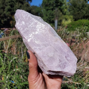 May include: A large, raw, pale pink crystal specimen with a jagged, elongated shape and a translucent, crystalline texture. The stone features natural facets and irregular edges, displaying a soft, light pink hue throughout its entire structure.