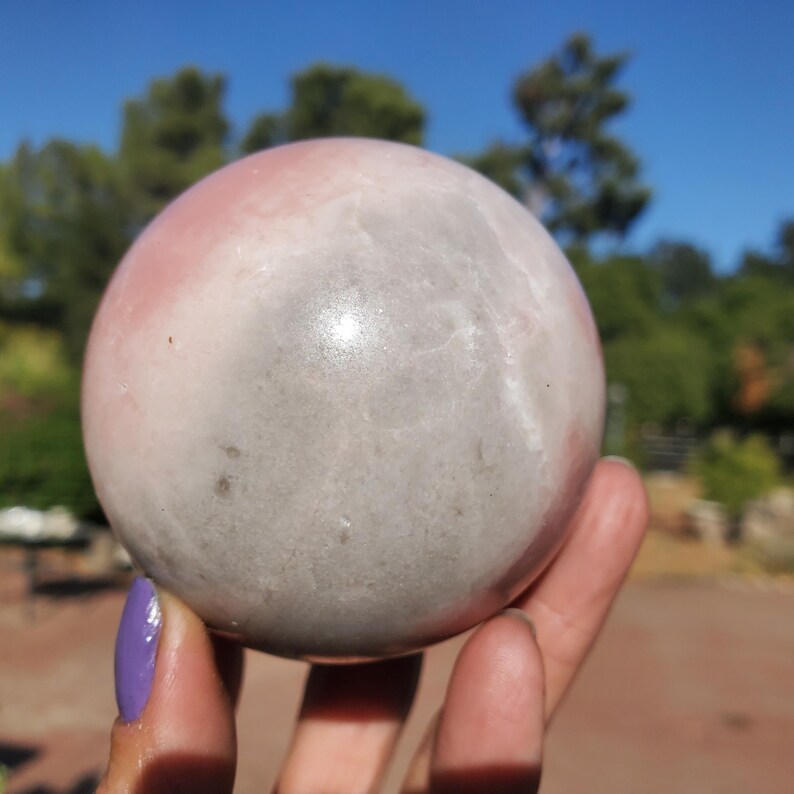 Peut inclure: Une pierre pr&eacute;cieuse sph&eacute;rique polie avec un d&eacute;grad&eacute; de teintes roses et grises. La pierre est tenue dans une main, avec un arri&egrave;re-plan flou d'arbres et un ciel bleu vif. La sph&egrave;re mesure environ 7.6 cm de diam&egrave;tre.