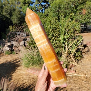 May include: A tall, hexagonal crystal tower made of orange calcite. The stone features a translucent, warm amber hue with distinct horizontal white banding and a smooth, polished finish that tapers to a sharp, faceted point at the top.