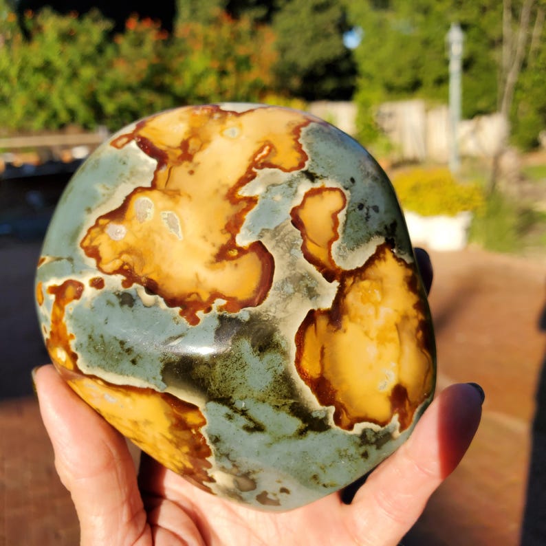 May include: A polished stone with a unique pattern of tan, brown, and blue-green colors. The stone has a smooth, rounded shape and appears to be a decorative item or a natural specimen. The colors and patterns resemble a map or landscape.