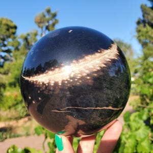 Può includere: Una sfera nera lucida con un motivo maculato marrone chiaro sulla sua superficie. La sfera è tenuta in mano su uno sfondo di fogliame verde e cielo blu. La superficie della sfera riflette la luce, evidenziando la sua consistenza liscia.