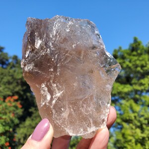 May include: A large, raw smoky quartz crystal held against a backdrop of a blue sky and green foliage. The crystal is a translucent brown color with a rough, uneven surface, showcasing natural formations. The light catches the facets, creating a shimmering effect.
