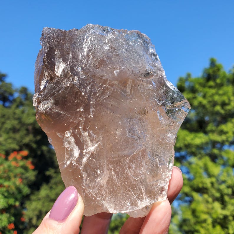 May include: A large, raw smoky quartz crystal held against a backdrop of a blue sky and green foliage. The crystal is a translucent brown color with a rough, uneven surface, showcasing natural formations. The light catches the facets, creating a shimmering effect.