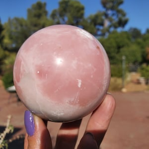 Peut inclure: Une grande sph&egrave;re de pierre pr&eacute;cieuse polie, dans les tons de rose et de blanc. L'orbe est pr&eacute;sent&eacute;e sur un fond d'arbres verts et de ciel bleu. La surface de la pierre a un aspect lisse et marbr&eacute;.