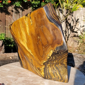 May include: A polished tiger's eye gemstone sculpture. The stone features swirling patterns of gold, brown, and black. The sculpture has a unique, asymmetrical shape and is displayed on a light-colored surface.