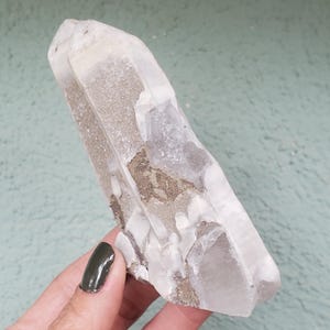 Pu&ograve; includere: Un cristallo di quarzo trasparente con una superficie ruvida e strutturata e una punta. Il cristallo presenta aree di inclusioni opache, marrone chiaro ed &egrave; tenuto su uno sfondo verde acqua chiaro. Il cristallo &egrave; lungo circa 13 cm.
