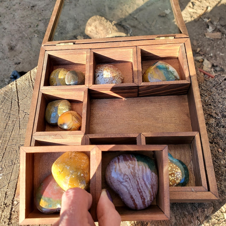 Op de afbeelding: Een houten doos met negen vakken, elk met een andere kleurrijke agaat steen. De doos is open en de stenen zijn zichtbaar.
