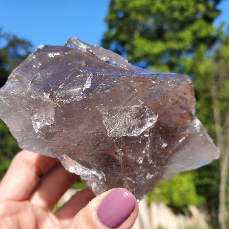 May include: A large, rough-cut smoky quartz crystal. The crystal is a translucent, brownish-gray color with sharp edges and a natural, unpolished appearance. The background is a blurred view of green trees and a blue sky.