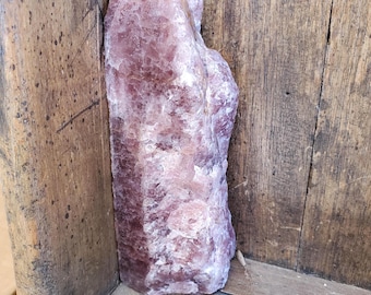 May include: A tall, raw, unpolished crystal specimen with a translucent, dusty rose and mauve coloration. The stone features a jagged, uneven surface with natural crystalline facets and a dense, semi-opaque texture throughout its vertical form.
