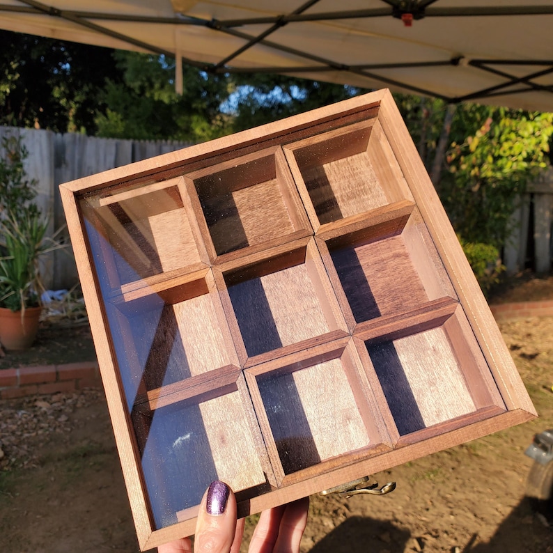 Op de afbeelding: Een houten doos met een glazen deksel en negen vakken. De doos is gemaakt van donker hout met een lichte houtnerf. De vakken zijn allemaal even groot en zijn gerangschikt in een 3x3 raster.