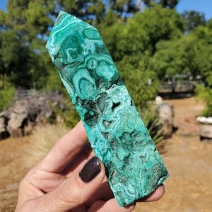 May include: A polished malachite crystal with a vibrant green color and intricate swirling patterns. The stone is rectangular with a flat top and a rough, natural edge. The crystal is held up against a blurred background of green trees and blue sky.