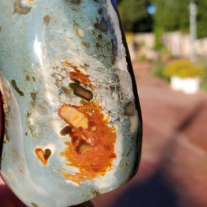 May include: A polished, irregularly shaped stone with a mix of colors. The stone features a light blue base with patches of brown, white, and dark green. A small heart-shaped indentation is visible on the surface.