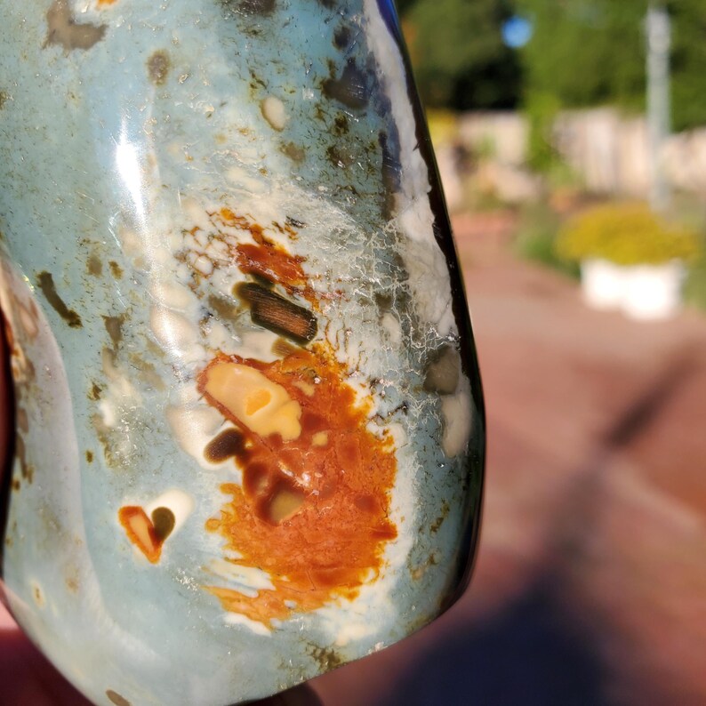 May include: A polished, irregularly shaped stone with a mix of colors. The stone features a light blue base with patches of brown, white, and dark green. A small heart-shaped indentation is visible on the surface.