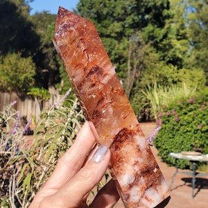 May include: A tall, six-sided crystal point featuring a blend of deep rust-red, burnt orange, and translucent white mineral inclusions. The polished stone displays a sharp, pointed apex and a thick, sturdy base with visible internal fractures.