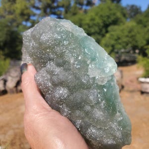 May include: A large, rough-hewn crystal formation with a mix of green and clear hues. The natural stone has a textured surface and is held in a hand, with a blurred outdoor background.