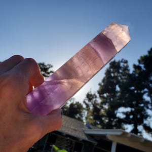 May include: A hand holds a large, polished crystal wand. The wand is a gradient of purple and clear, with visible internal patterns. The sunlight shines through the crystal, highlighting its translucent quality. The background features a blue sky and trees.