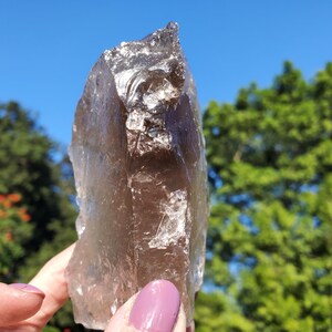 May include: A large, translucent smoky quartz crystal with a rough, natural texture. The crystal is a light brown color and is held against a backdrop of a blue sky and green foliage.